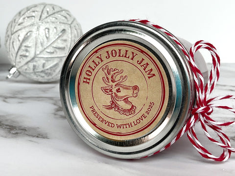 Round Kraft Apothecary Reindeer Christmas Canning Label on top of a mason jar lid. The label art is red with a  Rudolph the reindeer illustration in the center with text around the border reading Holly Jolly Jam | CanningCrafts.com