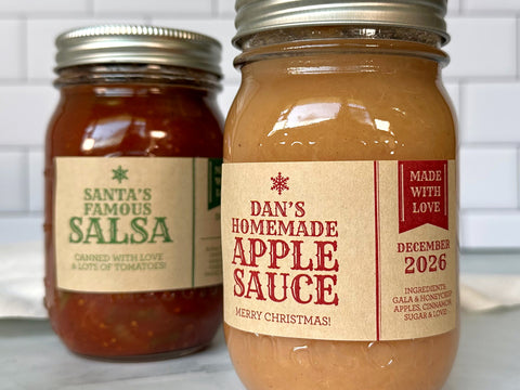 Two mason jars filled with food with rectangular kraft paper canning labels wrapped around them. The Apothecary Christmas Canning Labels are red and green with a small snowflake graphic. There is a cute pennant banner with text reading Made With Love. Each label is custom printed with text for the food inside such as applesauce and salsa. | CanningCrafts.com