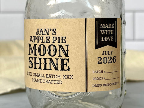 A mason jar filled with alcohol with a rectangular kraft paper canning label wrapped around it. The modern and rustic Apothecary Style Moonshine Label is black with a cute pennant banner with text reading Made With Love. Each label is custom printed with text for the type of alcohol such as moonshine, bourbon, or flavored liqueur. Blank lines can also be printed so text can be handwritten instead | CanningCrafts.com