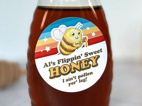 A Honey Bottle with round Custom Old School Honey Label on the front. The artwork shows a fuzzy retro honey bee holding a dipper on top of a colorful striped background with stars and custom printed text for beekeepers. Personalized honey label prints with your own custom text, such as “Flippin’ Sweet Honey, I Ain’t Pollen Your Leg”. | CanningCrafts.com