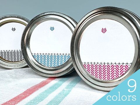 Three canning jar lids with our Blank Herringbone Canning Labels on the front. Each label has a colored pattern at the bottom with a small icon on the top. The colors are pink, blue, and black | Canningrafts.com